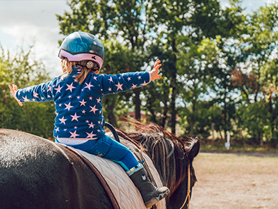 A young girl with a star-printed outfit and a helmet spreads her arms wide while riding a horse, depicting a sense of freedom and joy outdoors, embodying lessons learned from pet behavior guidance.
